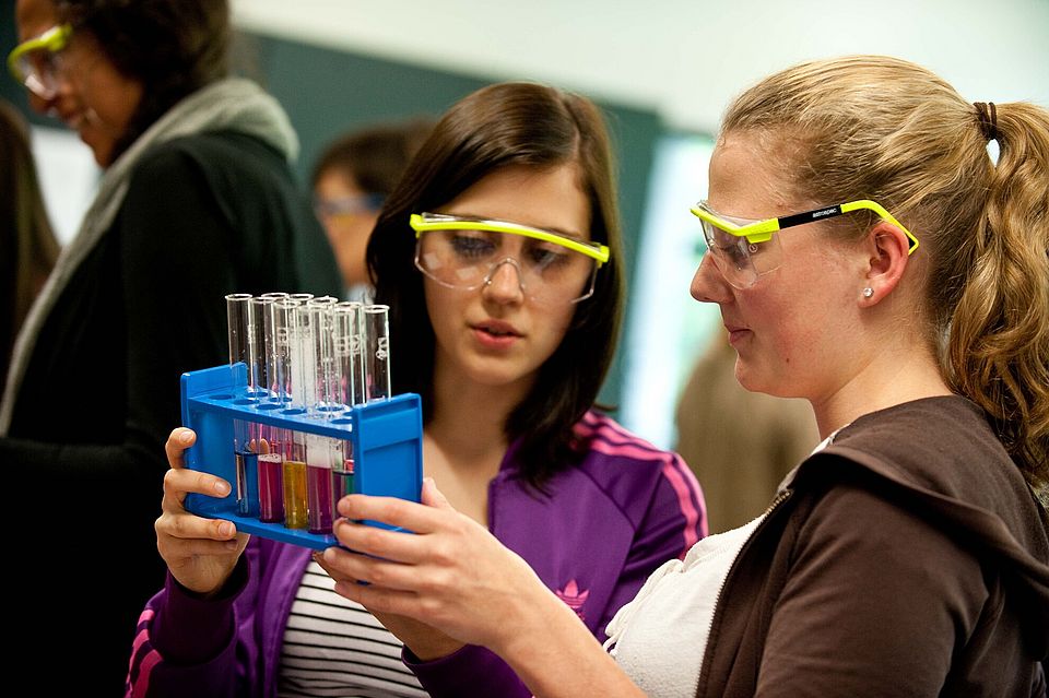 Zwei Schülerinnen mit Schutzbrillen und Reagenzgläsern in einer Halterung
